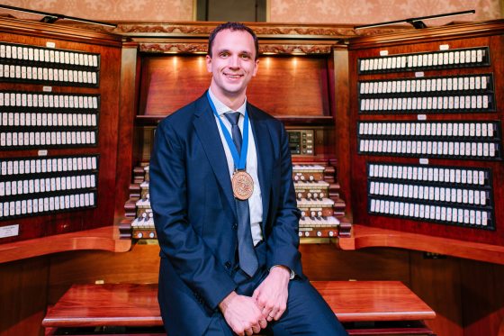 A person in a blue suit sitting at the bench in front of an organ console.